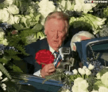 a man in a suit and tie is holding a red rose in front of a car filled with flowers