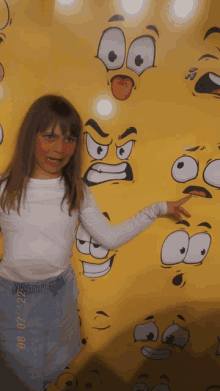 a young girl stands in front of a wall with cartoon faces painted on it