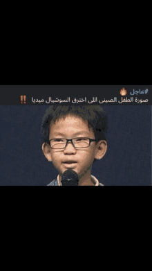 a young boy wearing glasses stands in front of a microphone with arabic writing below him