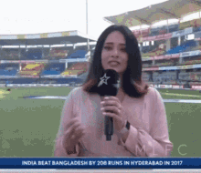 a woman is holding a microphone in front of a stadium that says india beat bangladesh by 208 runs in hyderabad in 2017