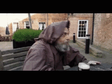 a man in a hooded robe sits at a table with a glass of beer in front of a brick building