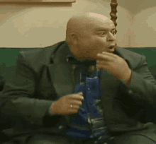 a bald man in a suit and tie is sitting on a couch talking on a cell phone .