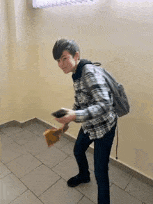 a young man with a backpack is standing on a tiled floor in a room holding a piece of paper .