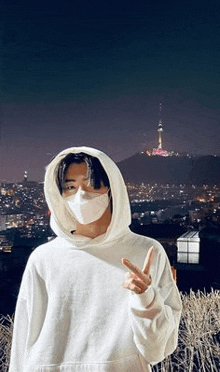 a young man wearing a white hoodie and a mask is standing in front of a city at night .