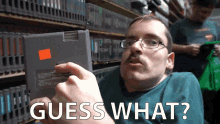 a man with glasses is holding a video game and says " guess what " in white letters