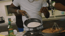 a man in a white shirt is cooking in a frying pan with a bottle of olive oil in the background