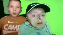 two boys are standing next to each other on a green screen . one boy is wearing a hat and headphones .