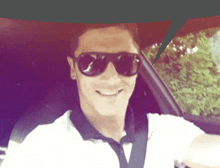 a man wearing sunglasses and a white shirt smiles while sitting in a car