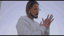 a man with a beard wearing sunglasses and a white sweater is standing with his hands folded in prayer .