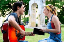 a man is playing a guitar to a woman in a park while she holds a book .