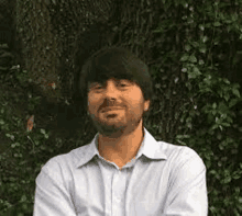 a man in a blue shirt is standing in front of a tree with his arms crossed .