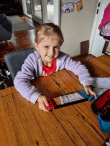 a little girl sits at a table with a nintendo switch in front of her