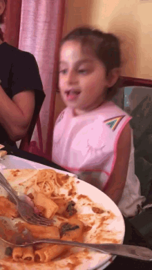 a little girl in a pink bib is sitting at a table with a plate of food and a fork