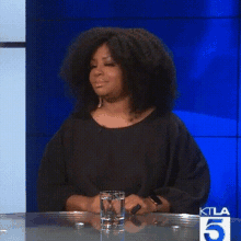 a woman is sitting at a table with a glass of water and a ktla 5 logo on the wall behind her .