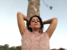 a woman in a pink lace top leans against a tree trunk