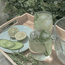 a wooden tray with two glasses of water with ice cubes and sliced limes and cucumbers