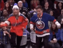 a man and a woman are dancing in the stands at a hockey game . the man is wearing a ny islanders jersey .