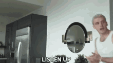 a man in a white tank top is standing in a kitchen next to a refrigerator and a mirror .