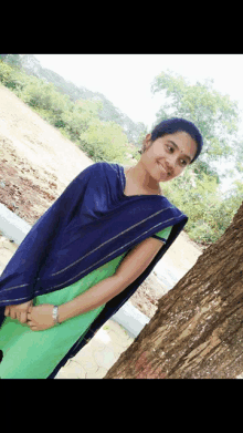 a woman in a green and blue dress is standing next to a tree and smiling