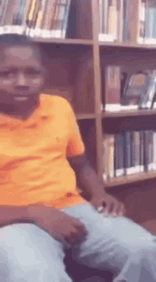 a man in an orange shirt and blue jeans is sitting in front of a bookshelf in a library .