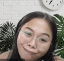 a woman wearing glasses is smiling in front of a clock and a plant .