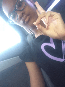 a girl wearing glasses and a black shirt with a purple heart on it giving the peace sign