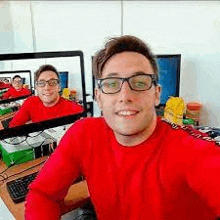 a man in a red sweater and glasses is taking a selfie in front of a computer screen .