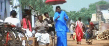 a man in a blue shirt is walking down a street with a red umbrella while people sit on benches .