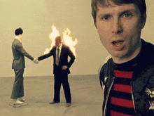 a man in a striped sweater stands in front of a man on fire