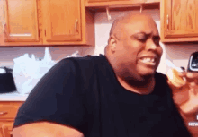 a man in a black shirt is eating a hamburger in a kitchen