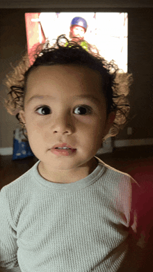 a young boy with curly hair looks at the camera in front of a tv screen