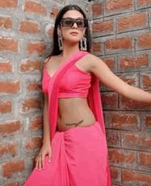 a woman wearing a pink saree and sunglasses is standing in front of a brick wall .