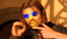 a man with a beard wearing blue sunglasses points at his mouth