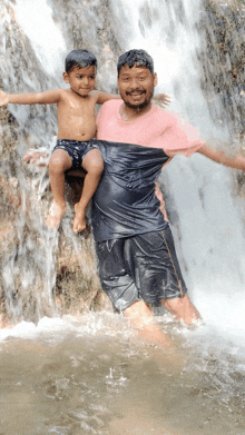 a man in a pink shirt is holding a little boy in his arms in front of a waterfall