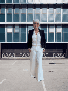 a woman wearing white pants and a black jacket stands in front of a large building