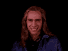 a man with long hair and a blue jacket is smiling in a dark room .