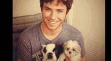 a man in a grey t-shirt with the letter t on it holds two puppies