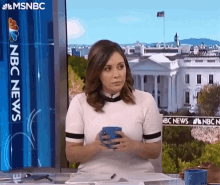 a woman is sitting at a desk holding a cup of coffee in front of a screen that says nbc news .