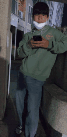 a young man wearing a green hoodie and a white mask holds a cell phone