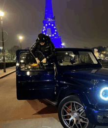 a man in a black jacket is sitting in a blue car in front of a blue eiffel tower