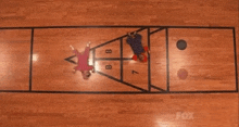 two people playing shuffleboard on a wooden floor with a fox logo in the corner