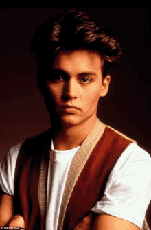 a young man wearing a brown vest and a white t-shirt is looking at the camera