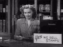 a black and white photo of a woman behind a desk with a sign that says yes we sell stamps