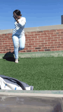 a woman in a white shirt and blue jeans is jumping in the air in front of a brick wall