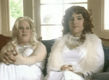 two women are sitting next to each other on a couch wearing white dresses