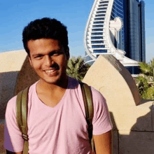 a young man wearing a pink t-shirt and a backpack is smiling in front of a building .