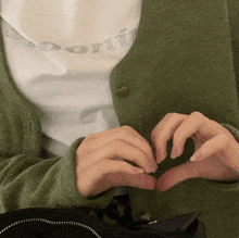 a person wearing a green cardigan making a heart with their fingers