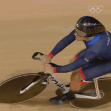 a man wearing a helmet and sunglasses is riding a bike on a track with the olympic rings in the background