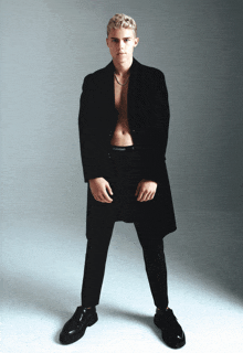 a shirtless man in a black coat and black calvin klein underwear stands with his hands on his hips