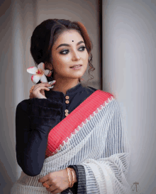 a woman wearing a black blouse and a white and red striped saree holds a flower